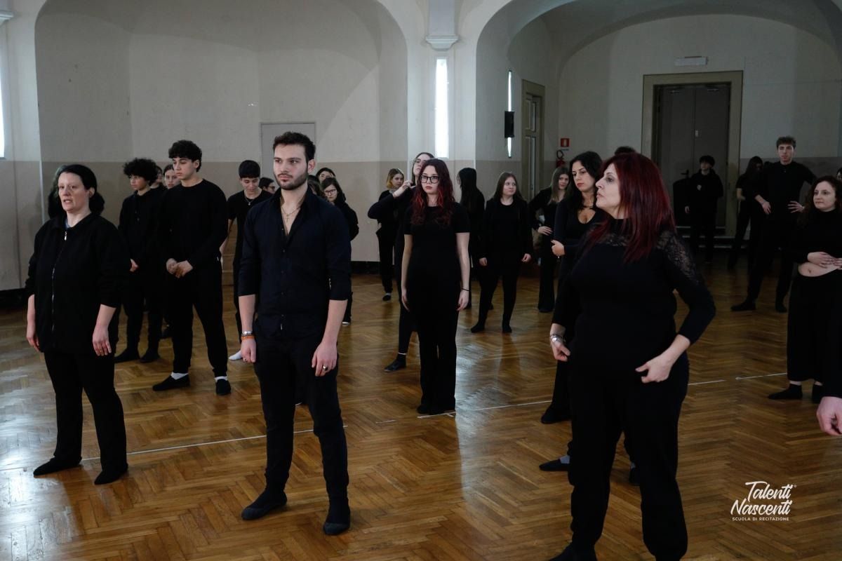 Un gruppo di persone vestite di nero è in piedi in una grande stanza, probabilmente impegnato a provare uno spettacolo.