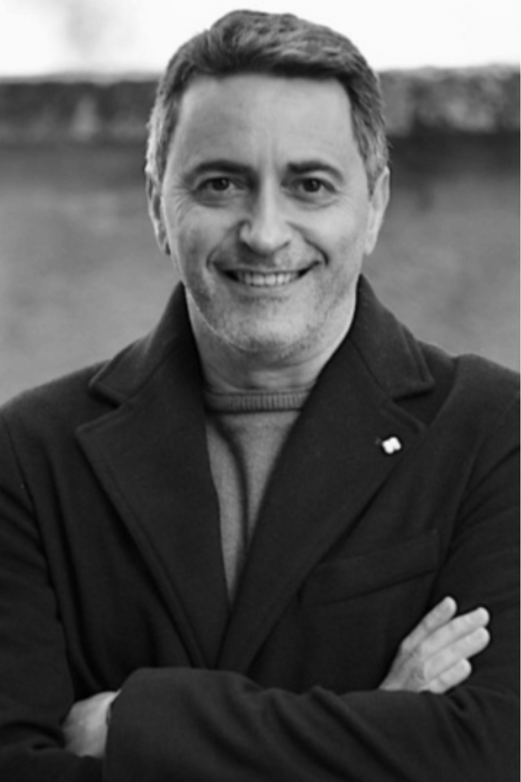 Un uomo con il cappotto sorride, braccia incrociate; all'aperto, con sfondo sfocato.