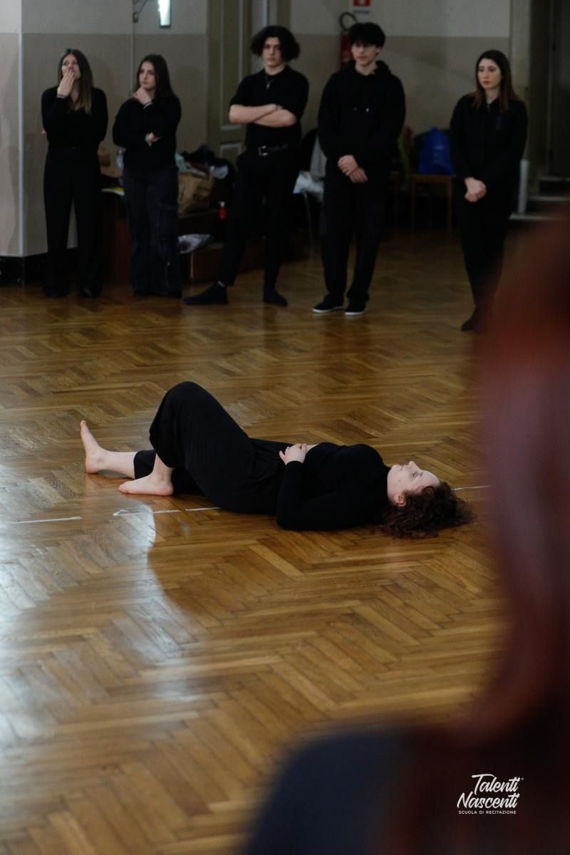 Una persona è sdraiata su un pavimento di legno e gesticola. Altri la osservano, vestiti di nero, in una stanza.