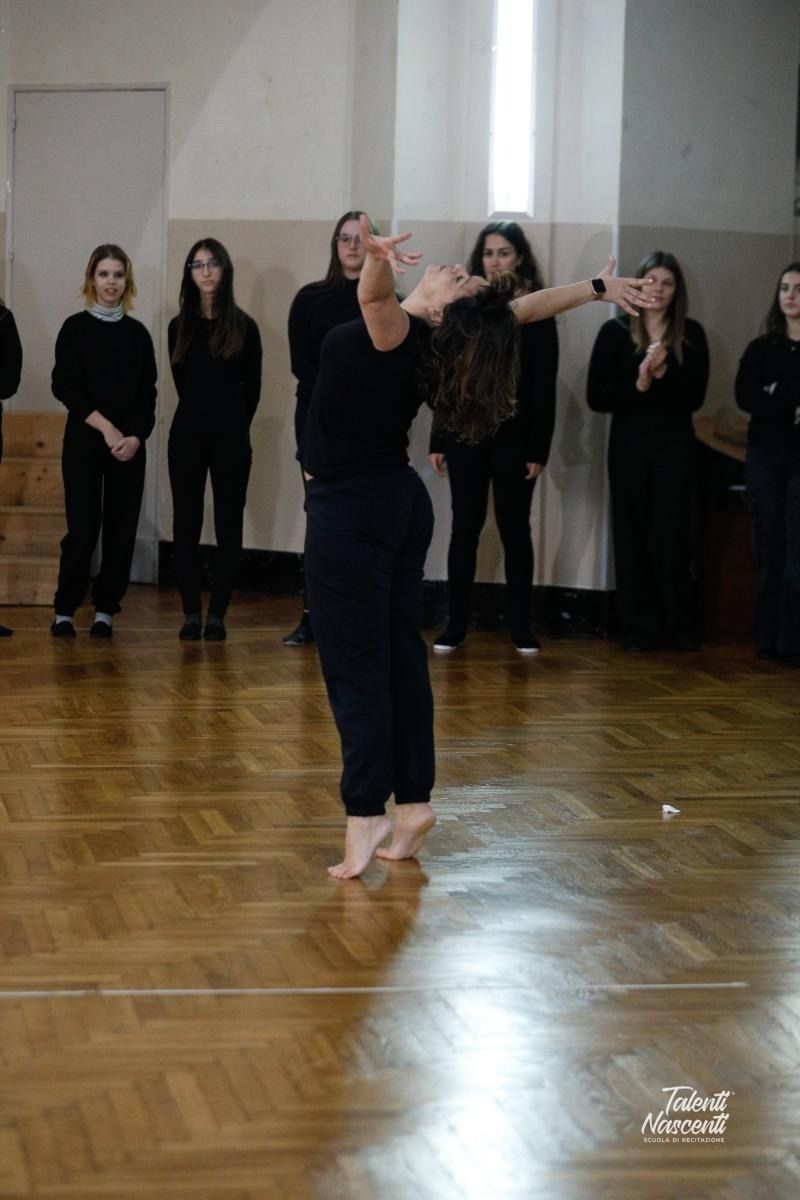 Una ballerina vestita di nero si esibisce su un pavimento di legno, sotto gli occhi di un gruppo di persone vestite di nero.