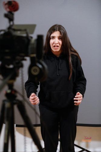 Una donna con felpa nera e pantaloni parla animatamente davanti a una telecamera montata su un treppiede.