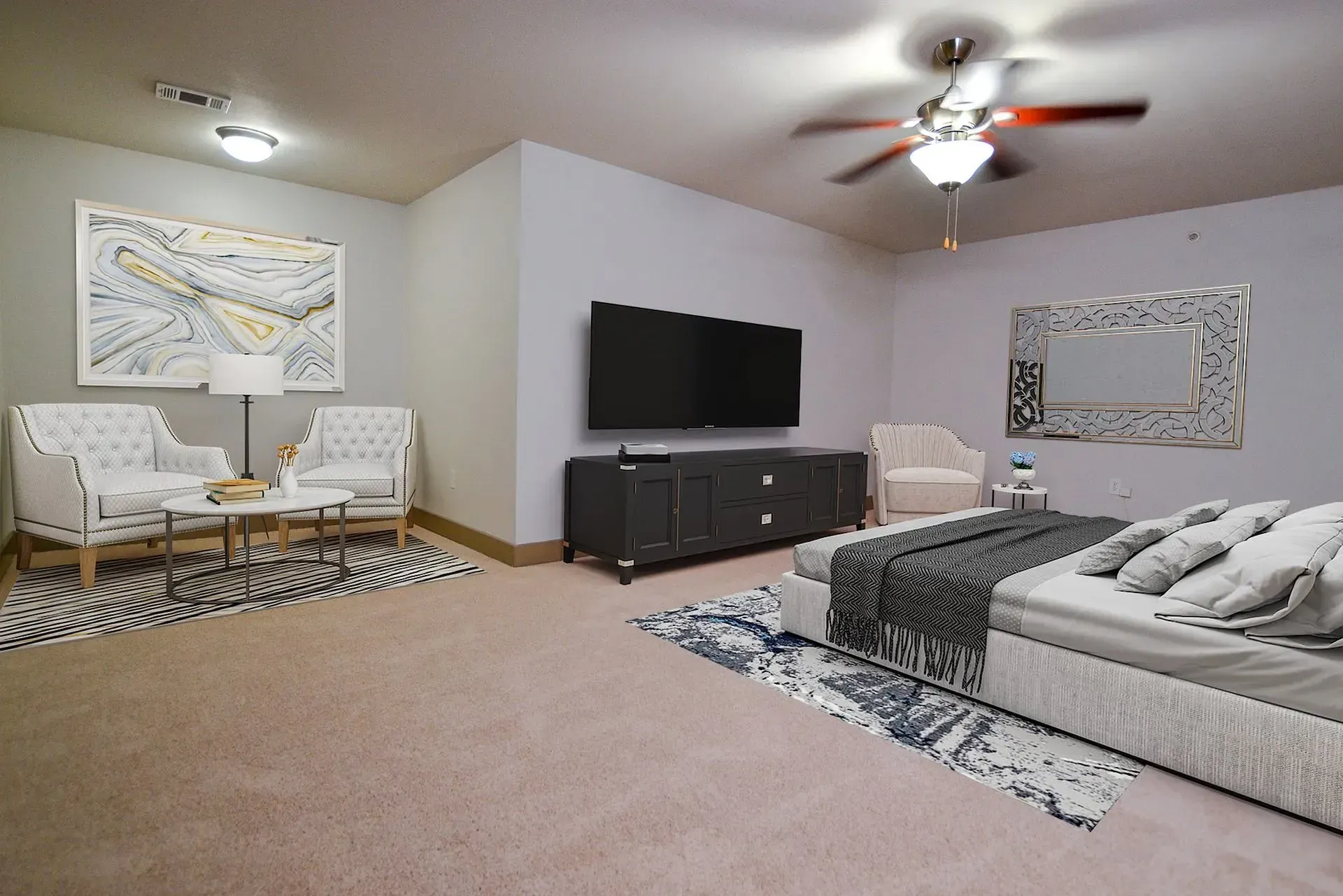 Spacious bedroom with a bed, seating area, TV, and a ceiling fan. Light-colored walls and neutral carpet.