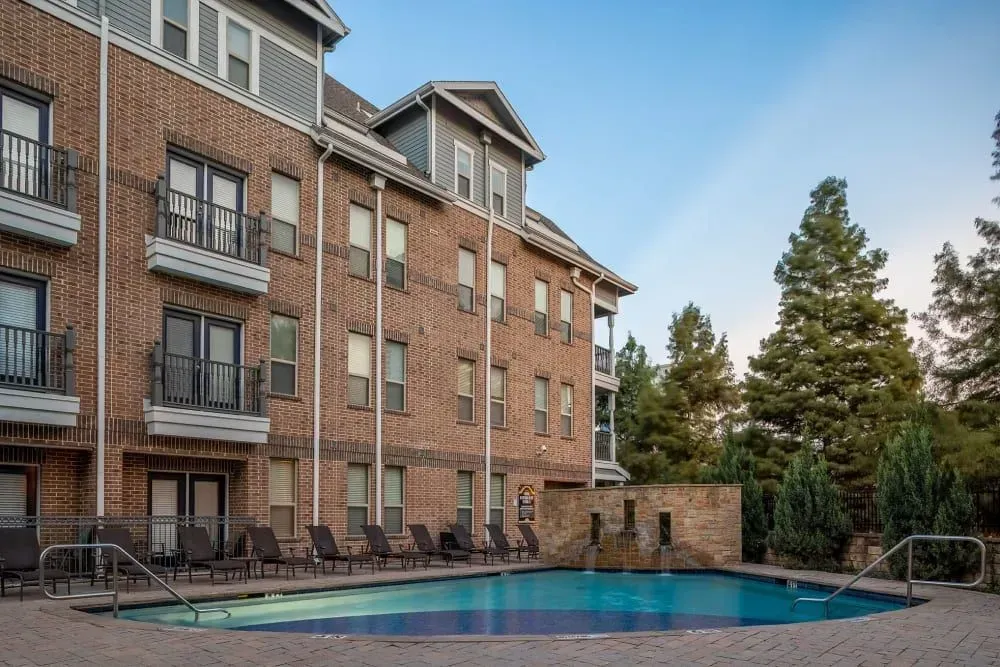 Apartment building with a pool, brick exterior, lounge chairs. Blue water, trees in the background.