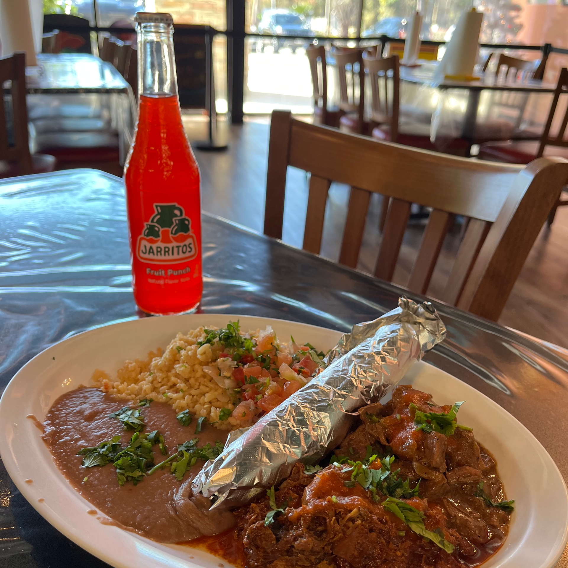 Plate of Mexican food, foil-wrapped item, refried beans, rice, Jarritos soda in a restaurant setting.
