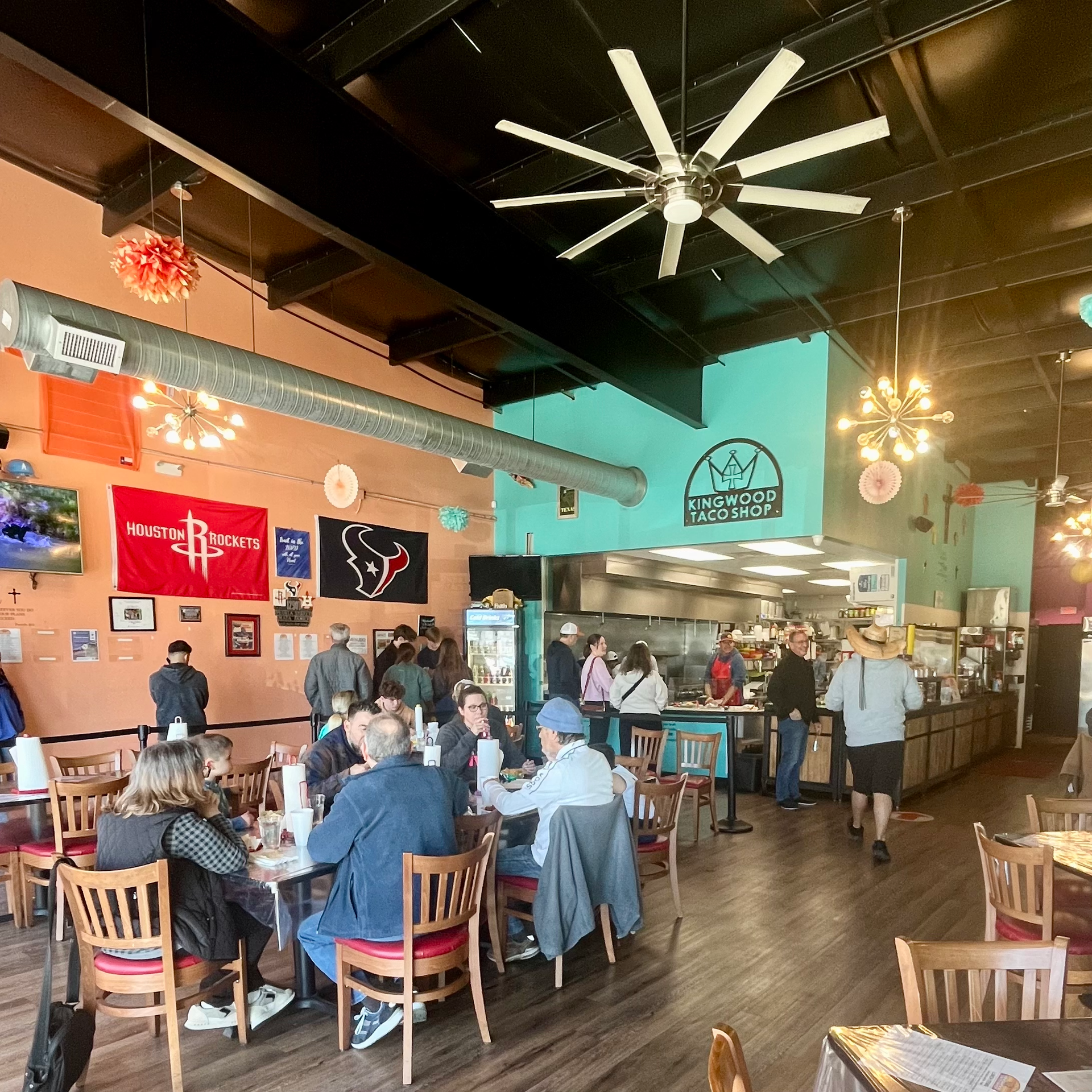 Interior of a restaurant with people dining and at a counter. Decorations include sports team flags and a large ceiling fan.