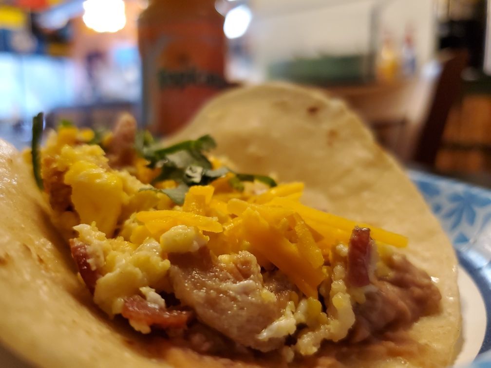 Taco filled with scrambled eggs, bacon, and cheddar cheese on a paper plate. A bottle of juice is in the background.