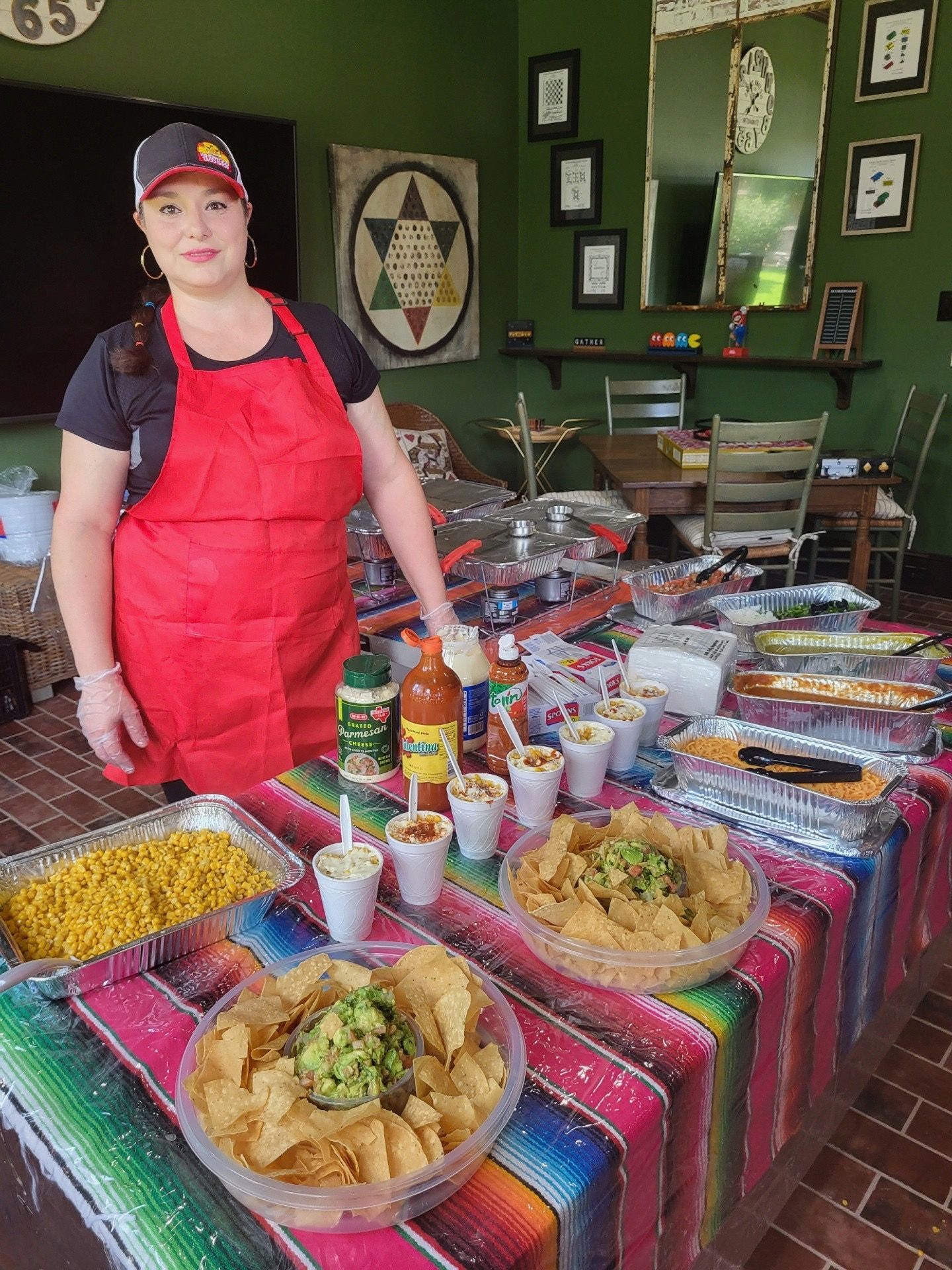 Woman in red apron stands behind a table of Mexican food. Colorful serape tablecloth, chips, guacamole, and sauces.