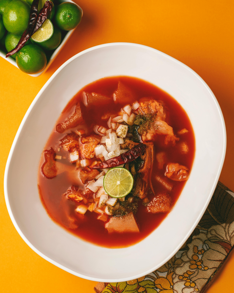 A bowl of soup with meat , onions and limes on a table.