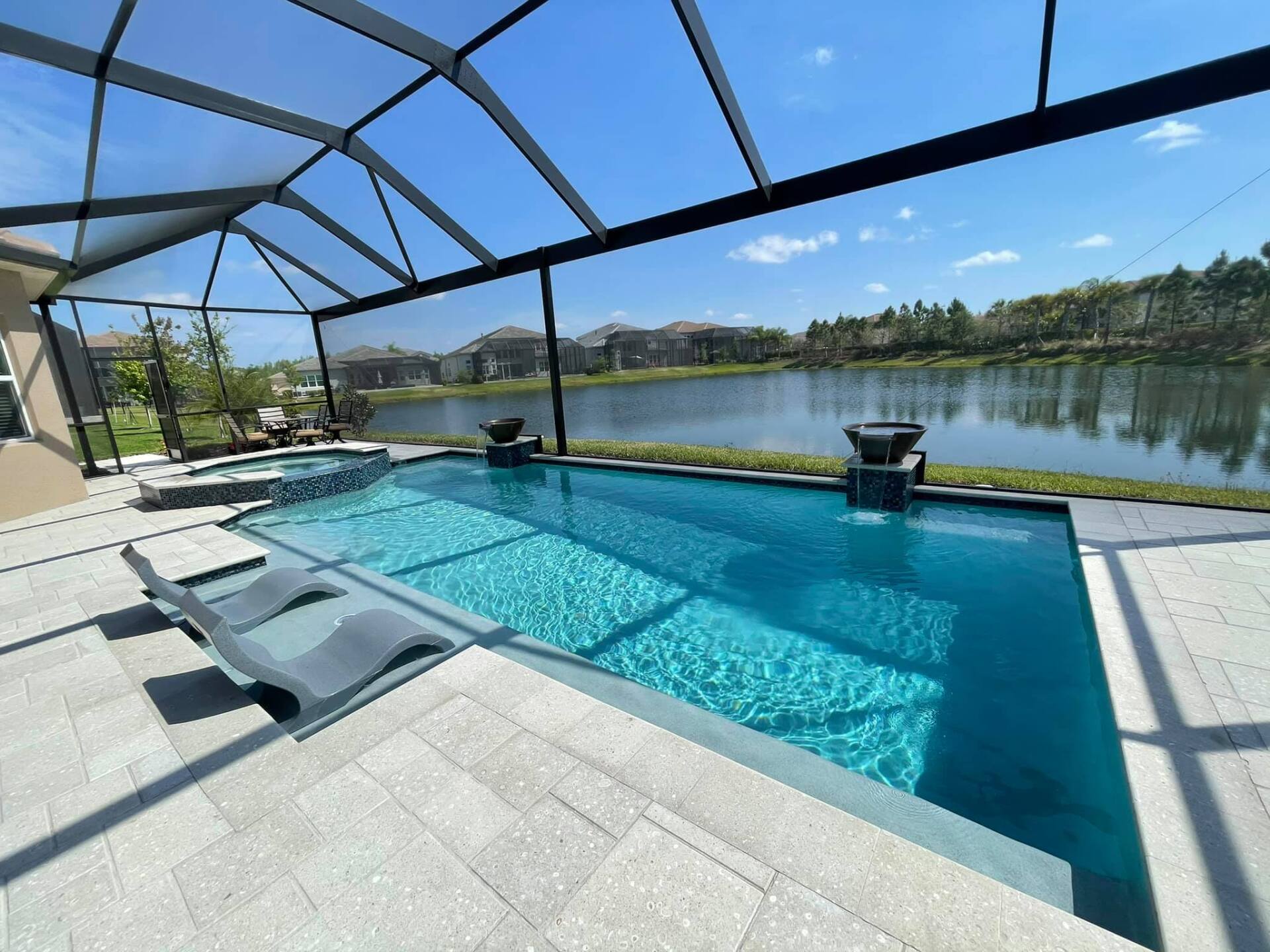 Pool with clear blue water, lounge chairs, and a screened enclosure overlooking a lake on a sunny day.