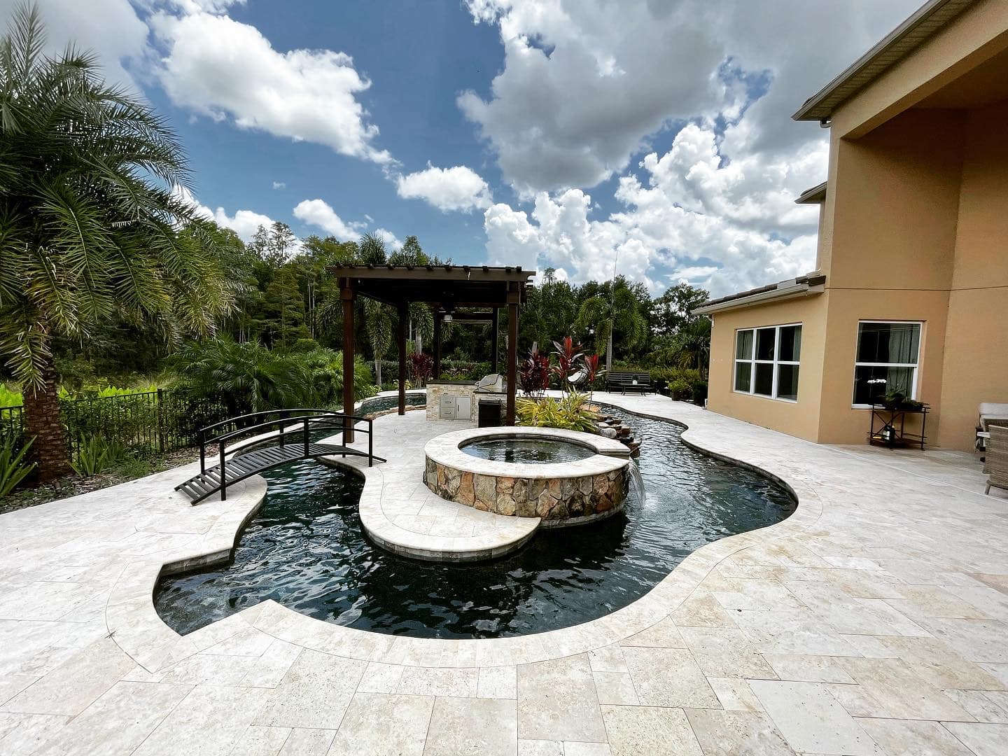 Backyard pool with travertine patio, fire pit, and pergola under a bright blue sky.