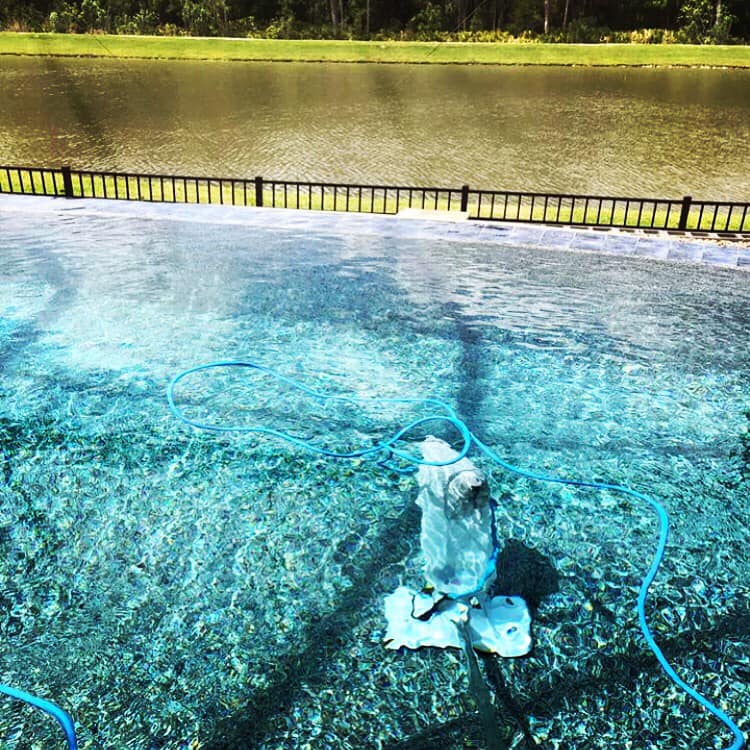 A robotic pool cleaner in a clear, blue pool near a fence and a lake.