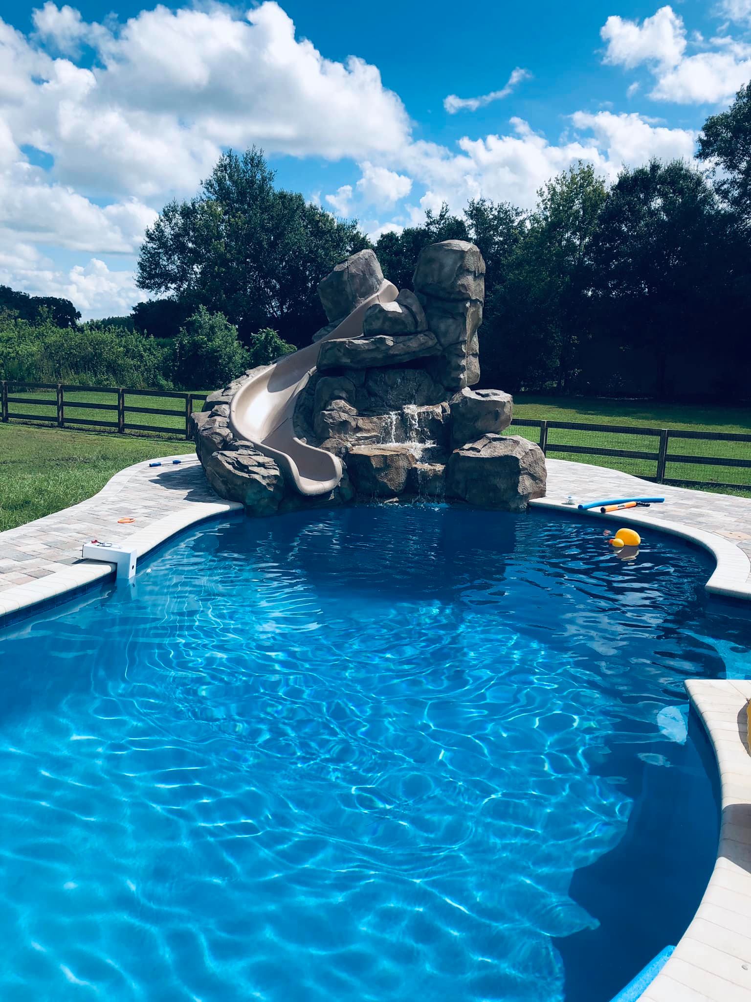 Swimming pool with rock waterfall slide under a bright blue sky.