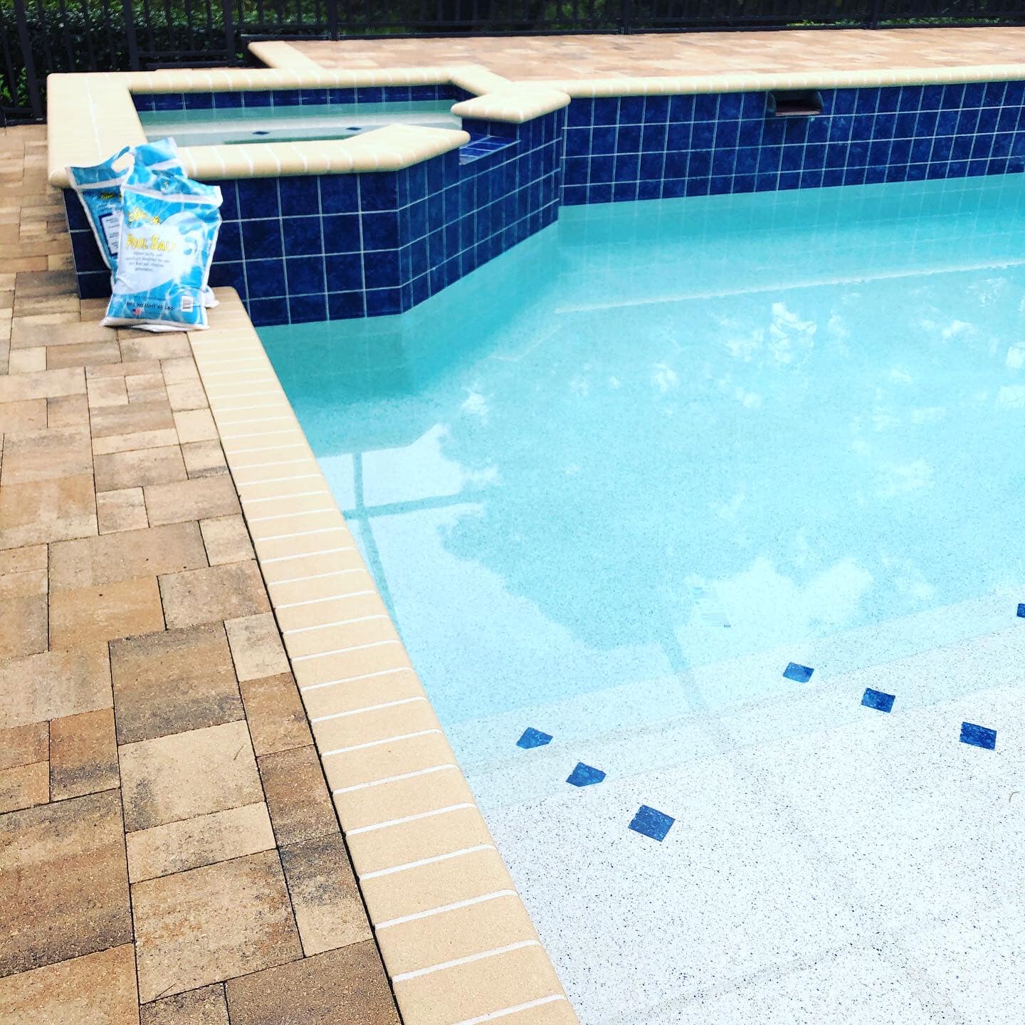 A pool with blue tiles and a tan brick patio. Two bags of pool chemicals sit near the pool.