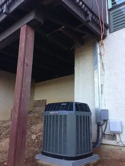 A large air conditioner is sitting under a deck next to a building.