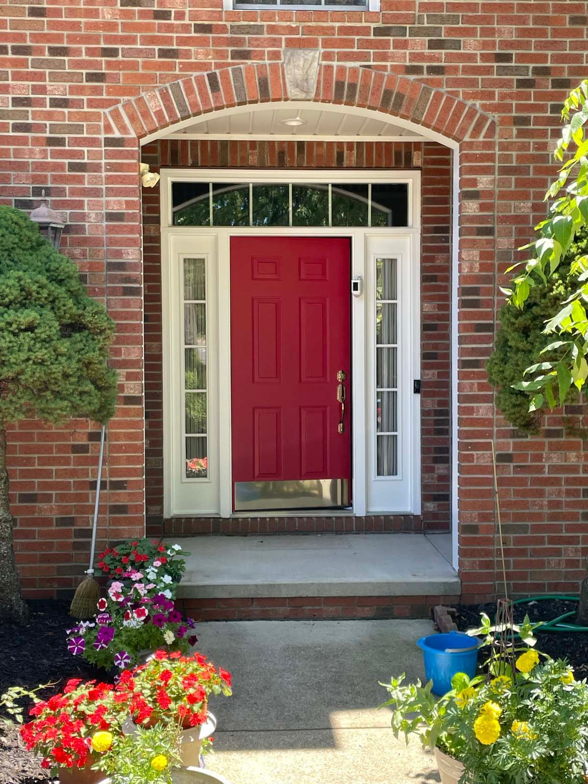 Brick house entry with a red front door, white trim, and colorful flowers by the walkway