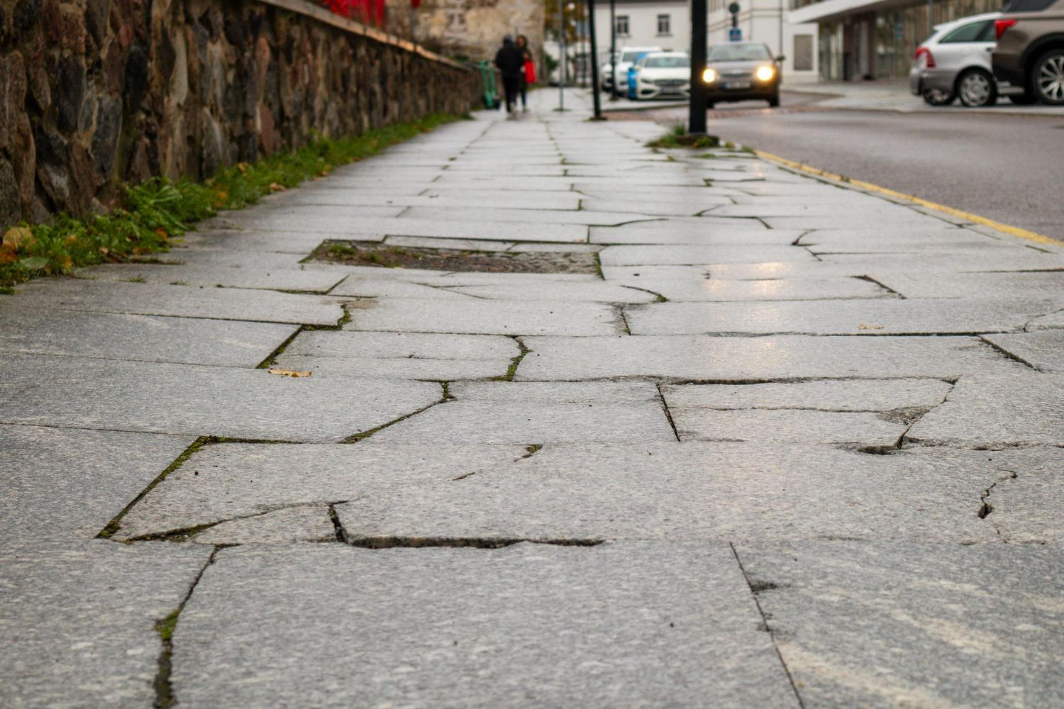 Cracked gray sidewalk with uneven stones next to a stone wall and street. A person and car are visible.