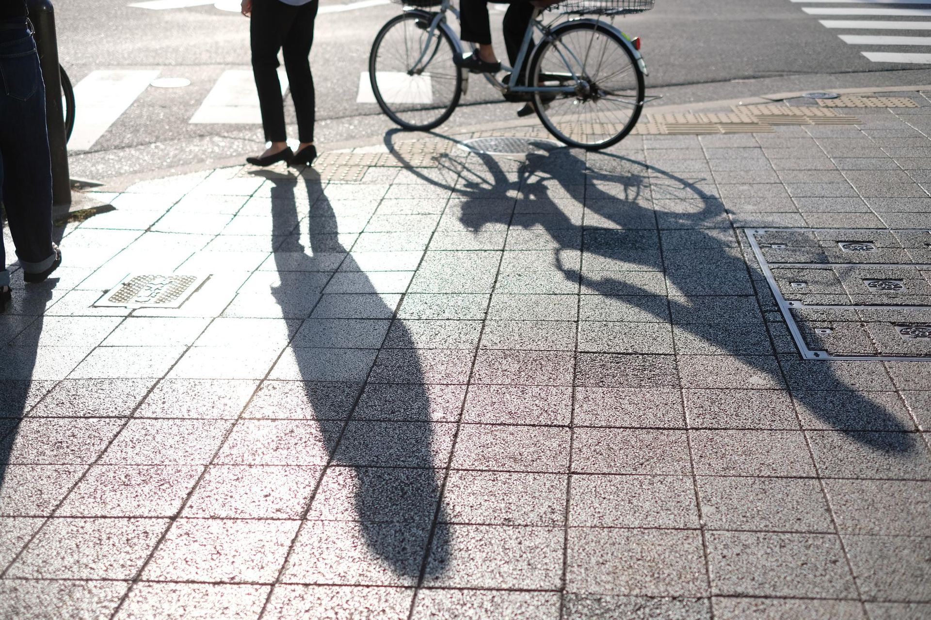 Shadow of person on a bicycle cast on a sunny sidewalk, with pedestrian in the left.