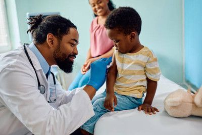 A Black doctor gives a vaccine to a child as the mother watches in a medical office.