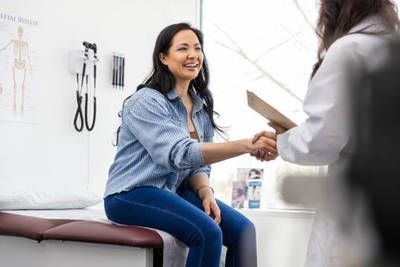Woman in jeans and shirt shakes hands with a doctor in an exam room.