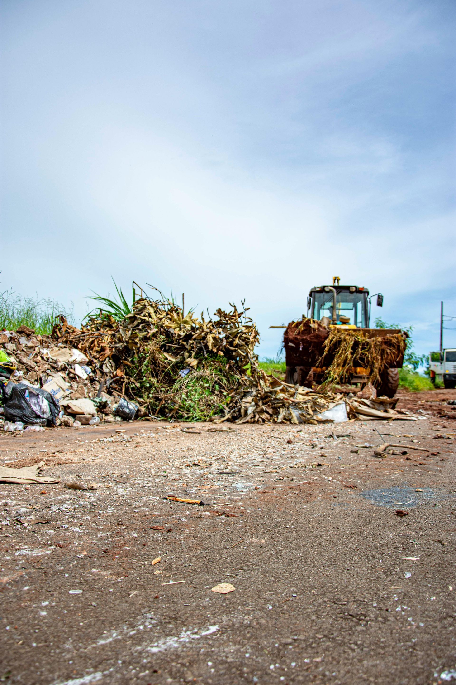 A tractor moves a large pile of yard waste and debris on an unpaved, dirt surface under a cloudy sky.