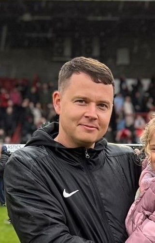 A man in a nike jacket is holding a little girl on a soccer field.