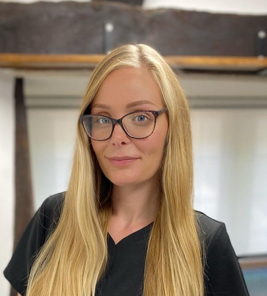 A woman with long blonde hair and glasses is wearing a black scrub top.