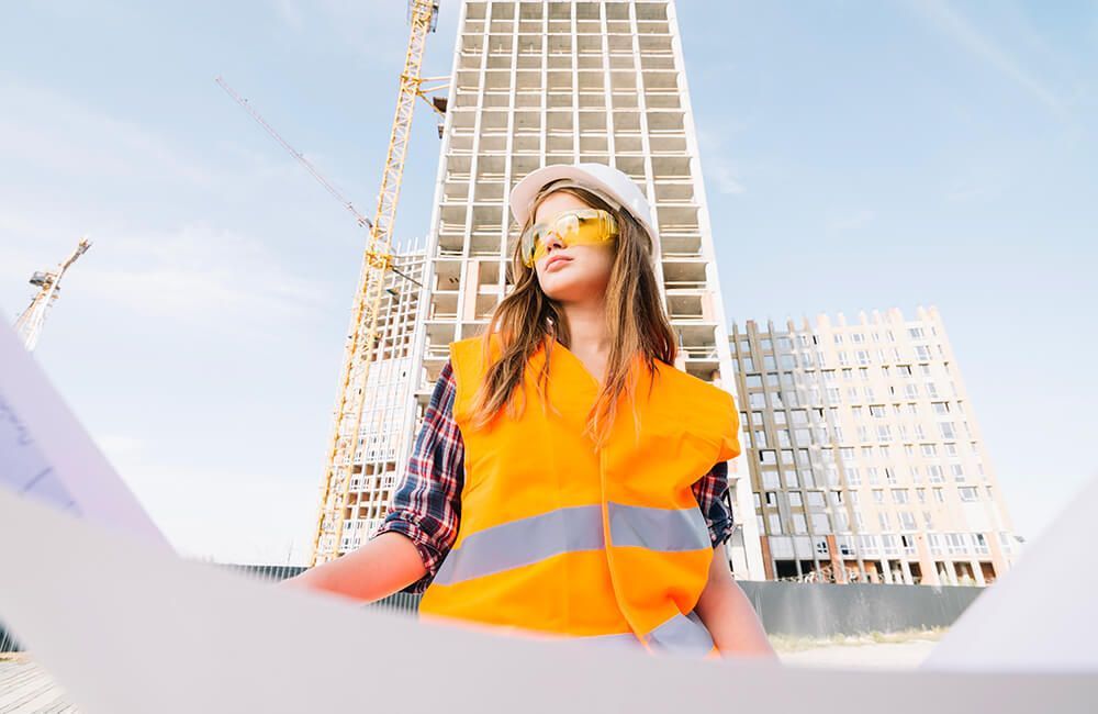 A woman is standing in front of a building under construction holding a blueprint.