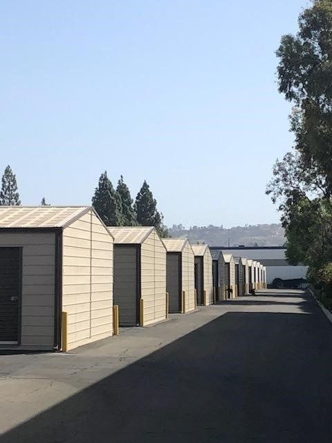Row of beige storage units with dark doors and tan trim, asphalt drive.