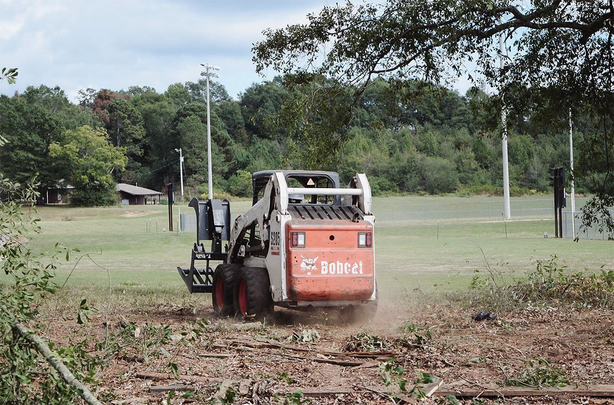 Bobcat skid-steer clears brush from a grassy field. Orange and white machinery, trees in background, sunny day.