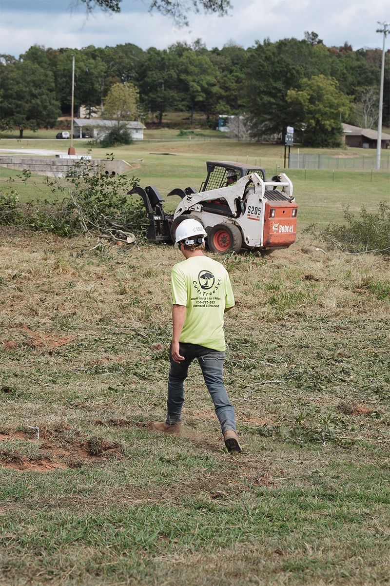 Person in a hard hat and neon shirt watches a Bobcat skid-steer loader clearing brush in a grassy field.