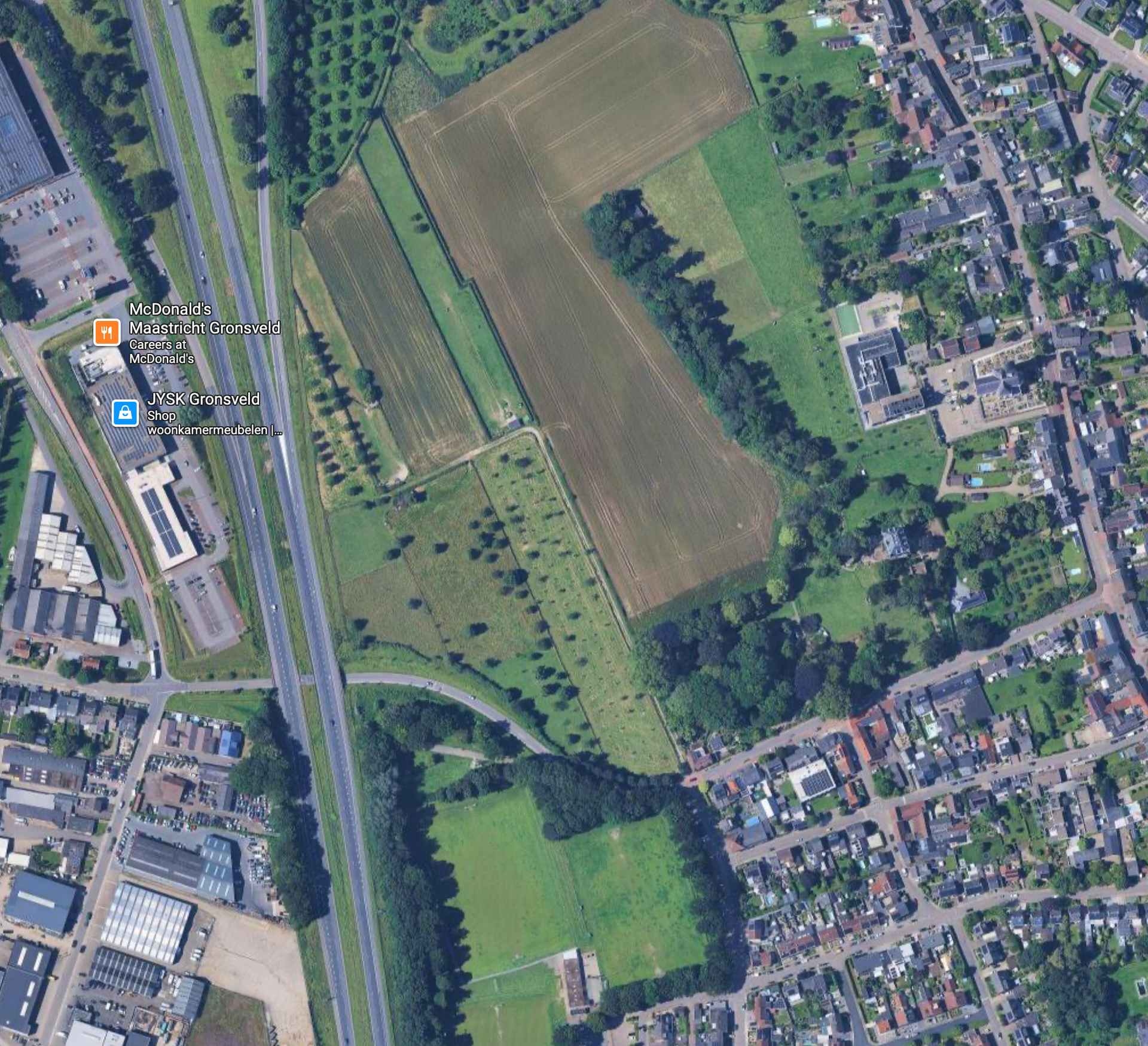 Luchtfoto van een veld, omgeven door bomen en een snelweg, met gebouwen aan de linkerkant en een woonwijk aan de rechterkant.