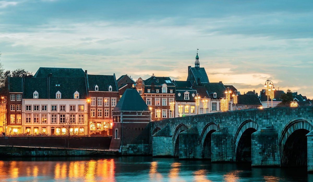 Stenen brug over het water, gebouwen met verlichte ramen, een kerktoren op de achtergrond in de schemering. Maastricht