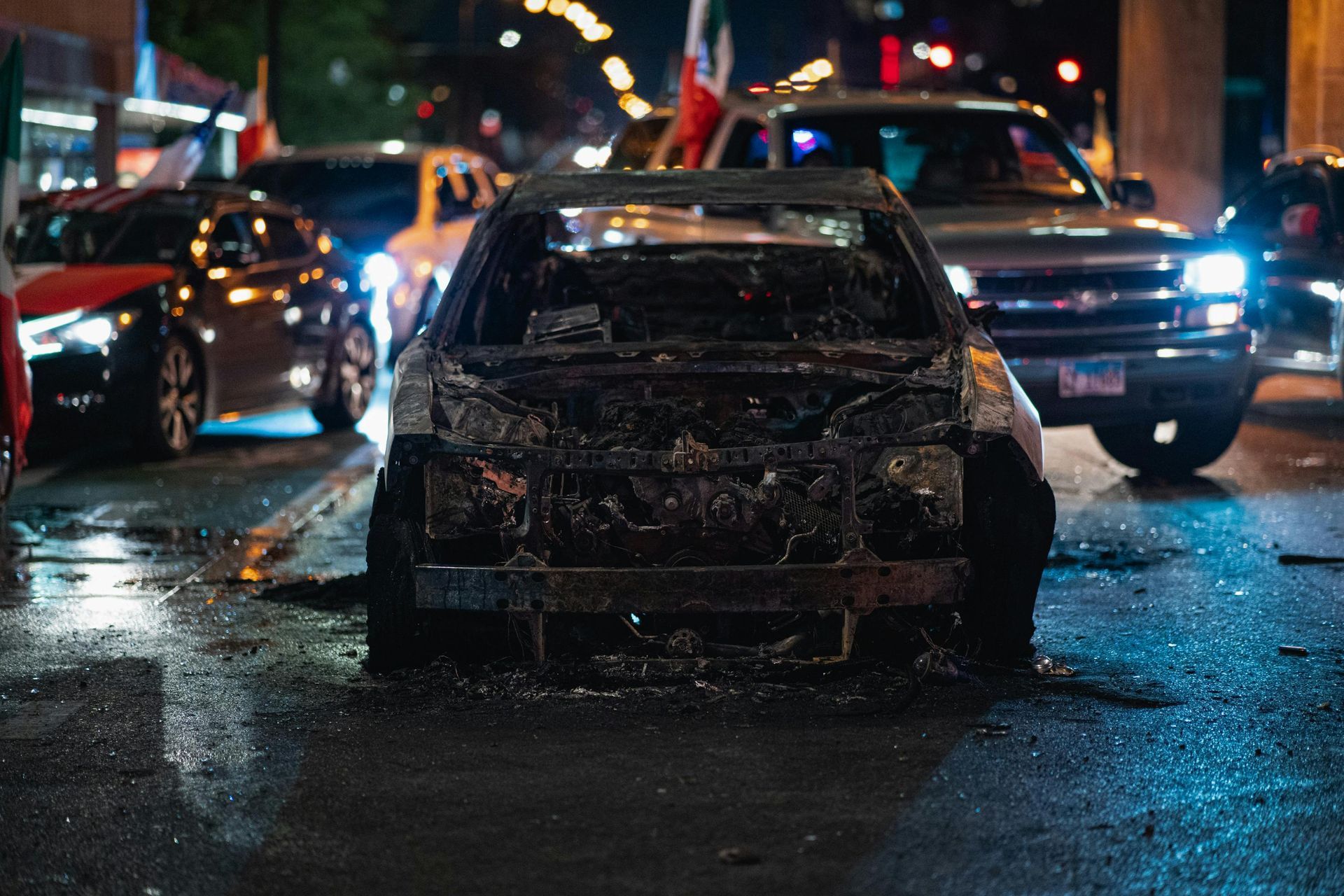 Burnt-out car on a wet city street at night, other vehicles and flags visible in the background.