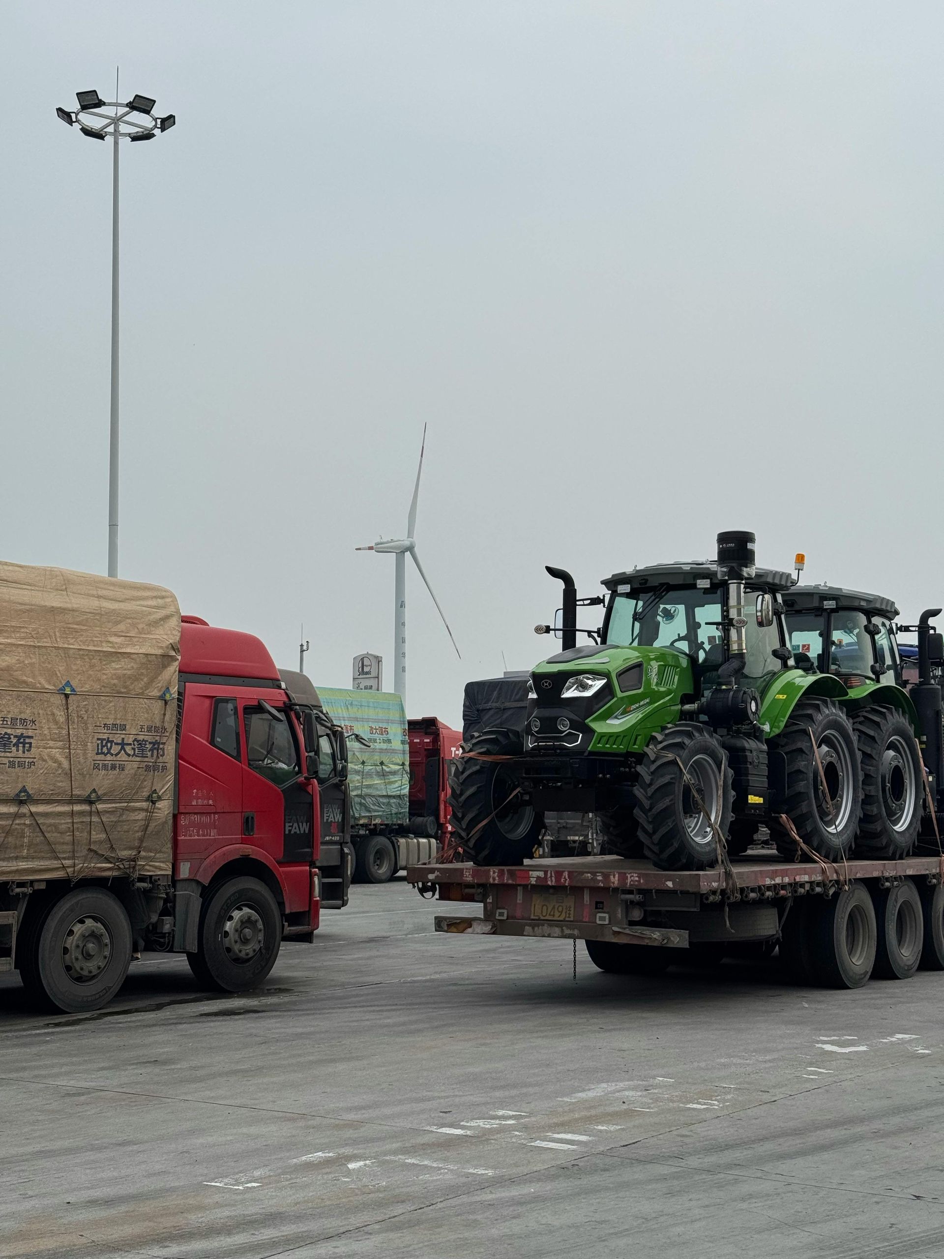 Red truck next to a flatbed with three green tractors. A wind turbine and a light pole are in the background.