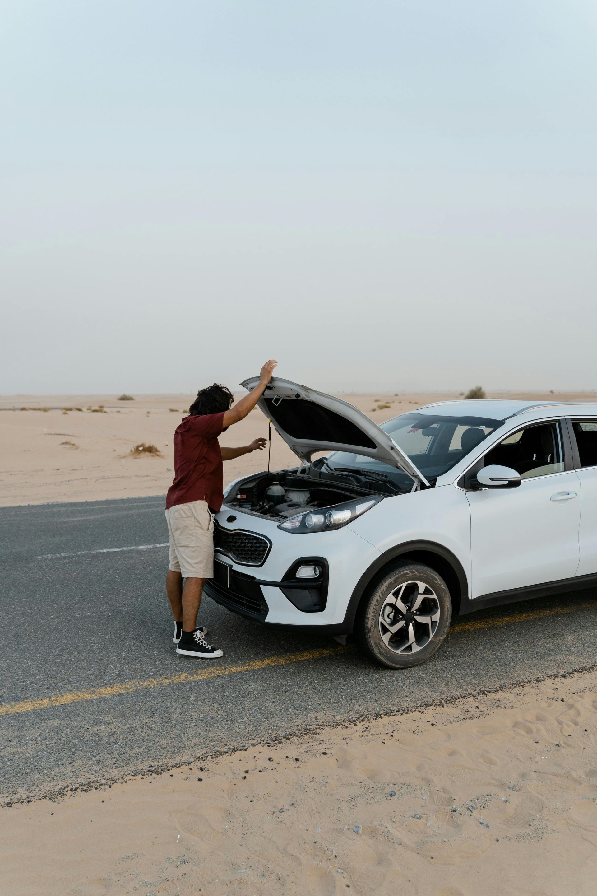 Man in shorts looking at a white car with its hood open on a desert road.