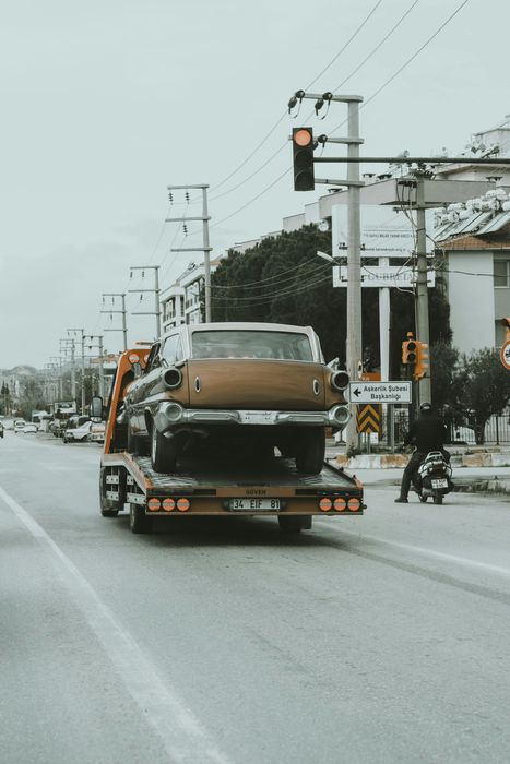 Classic car being towed on a flatbed truck on a city street. Grey sky, traffic light.
