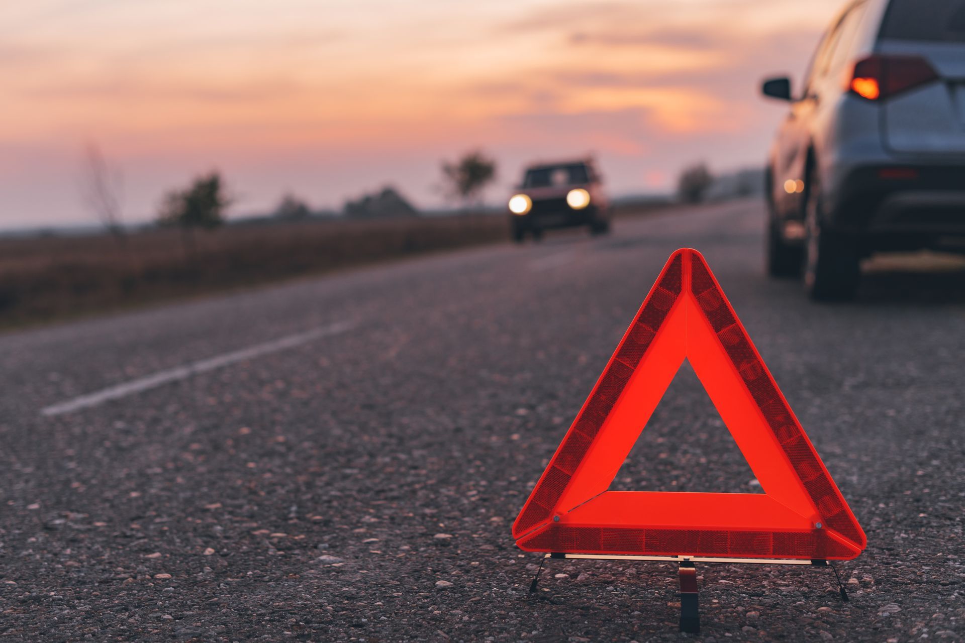Red warning triangle on asphalt road with a car, sunset in the background.