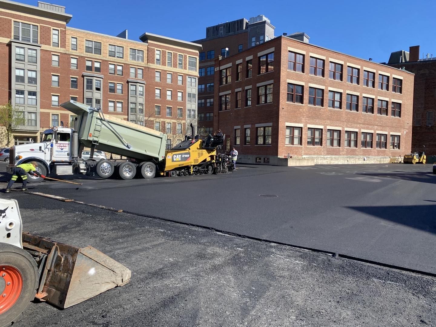 A dump truck is being loaded with asphalt in front of a brick building.