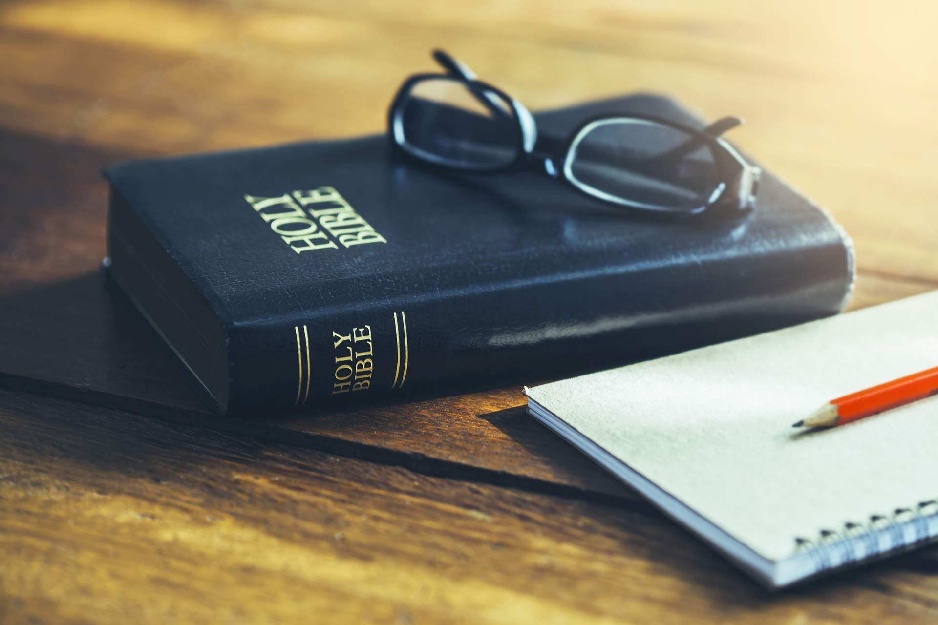 A bible , glasses , a notebook and a pencil are on a wooden table.
