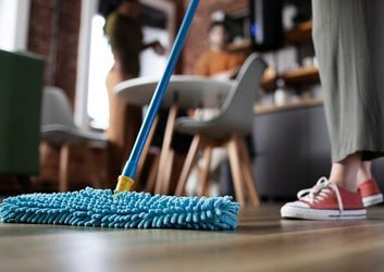 A person is standing next to a broom and shovel on a wooden floor.