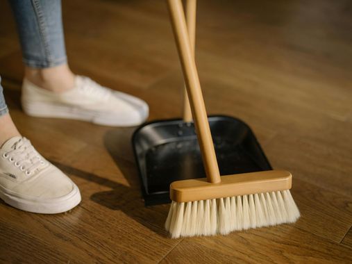A person is standing next to a broom and shovel on a wooden floor.