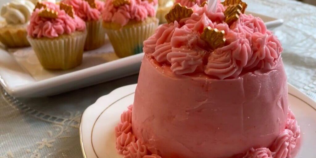 A small pink cake is on a plate next to a plate of cupcakes.