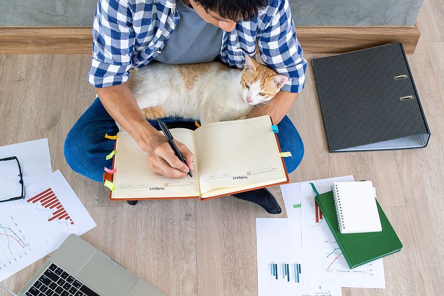 A man is sitting on the floor with a cat on his lap while writing in a notebook.