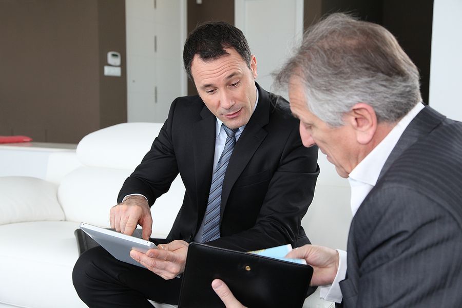 Two men in suits are sitting on a couch looking at a tablet.