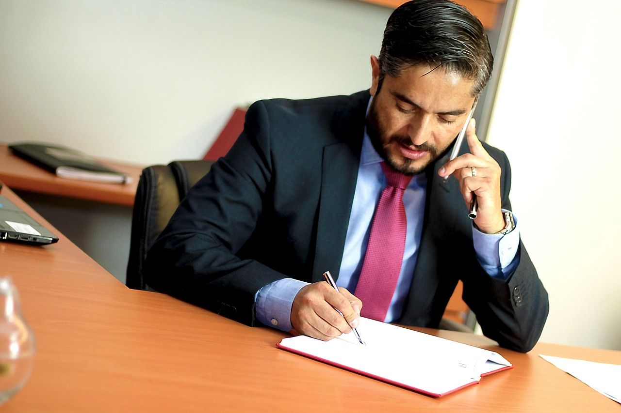 A man in a suit and tie is talking on a cell phone while writing on a clipboard.