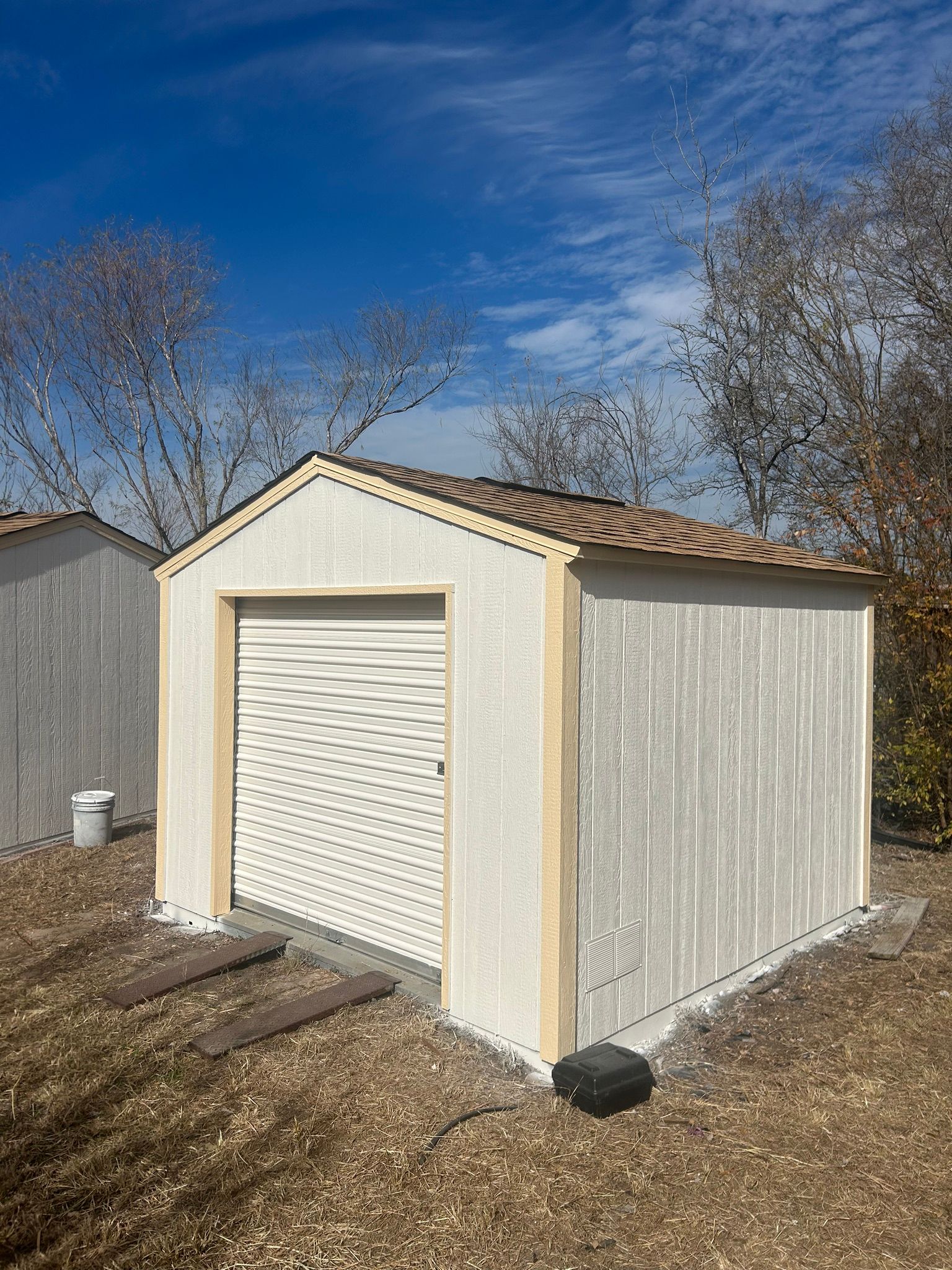 A small white shed with a brown roof is sitting in the middle of a dirt field.