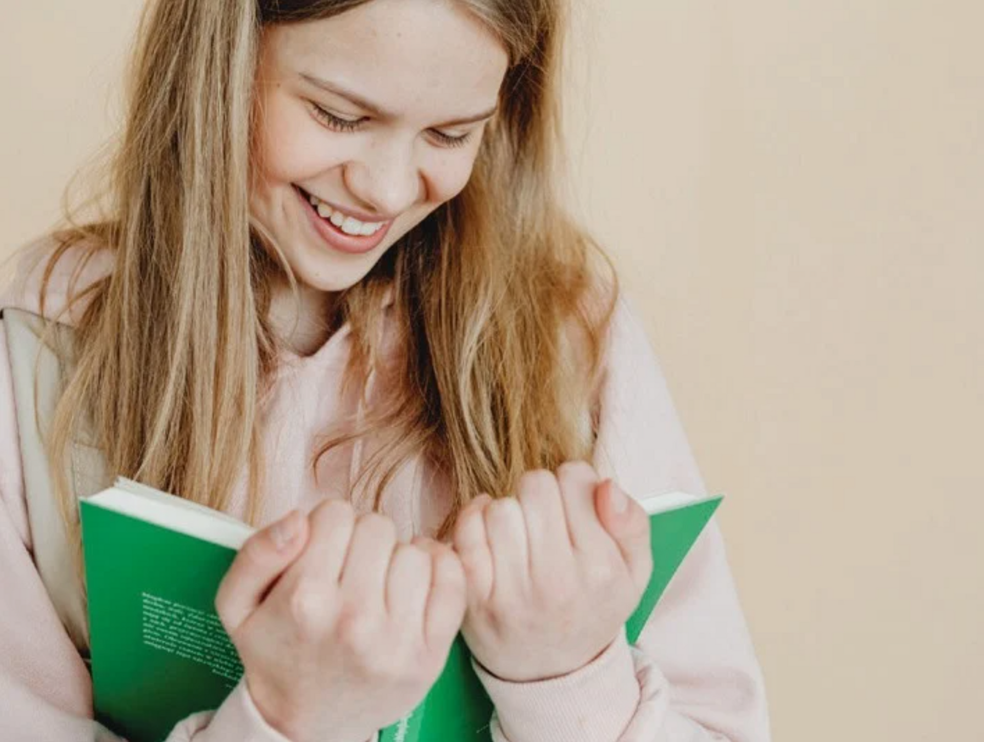 Blonde person smiling while holding a green book, wearing a pink hoodie against a neutral background.