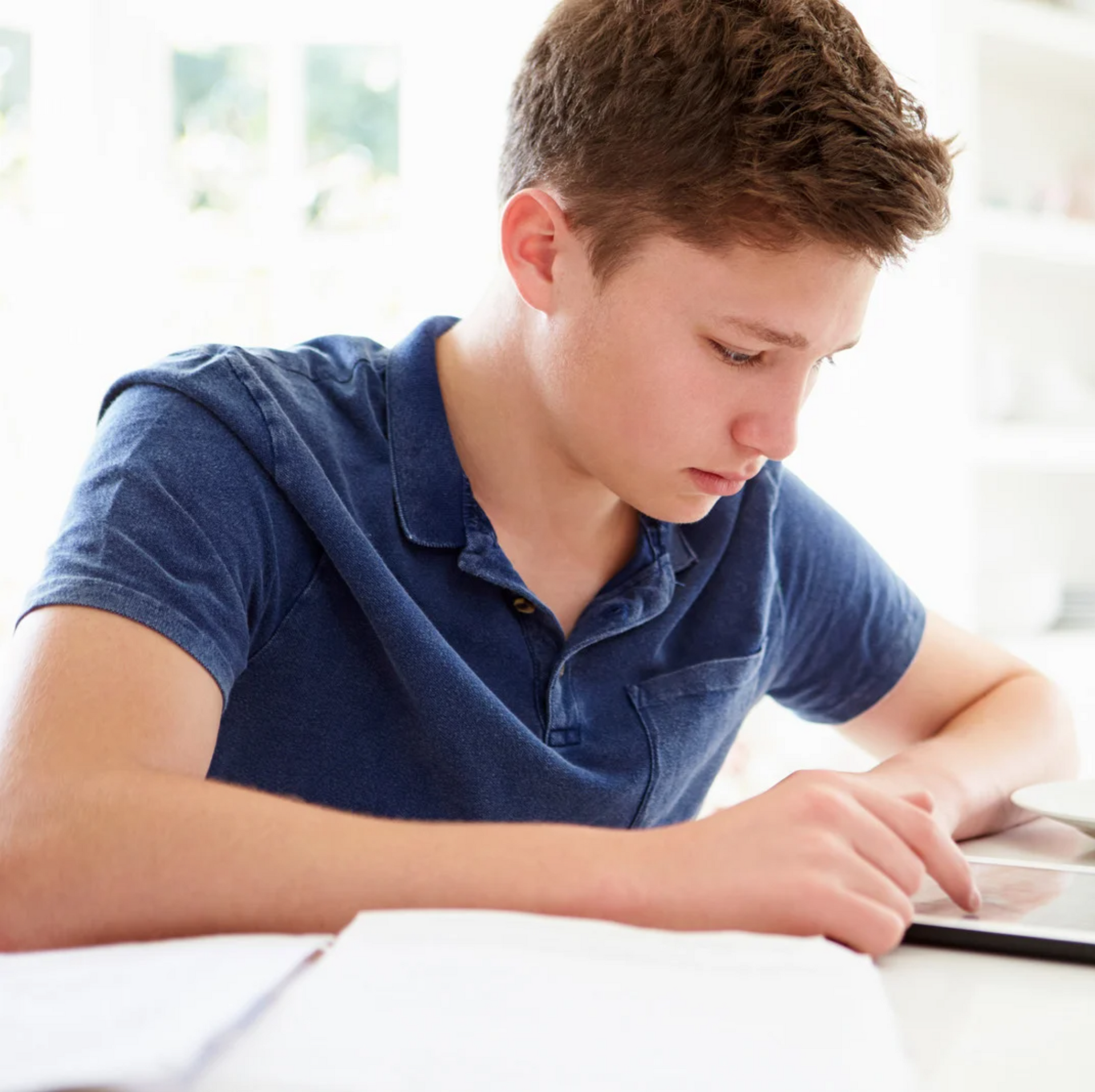 Teenager in blue shirt focused on tablet, seated at a desk with papers.