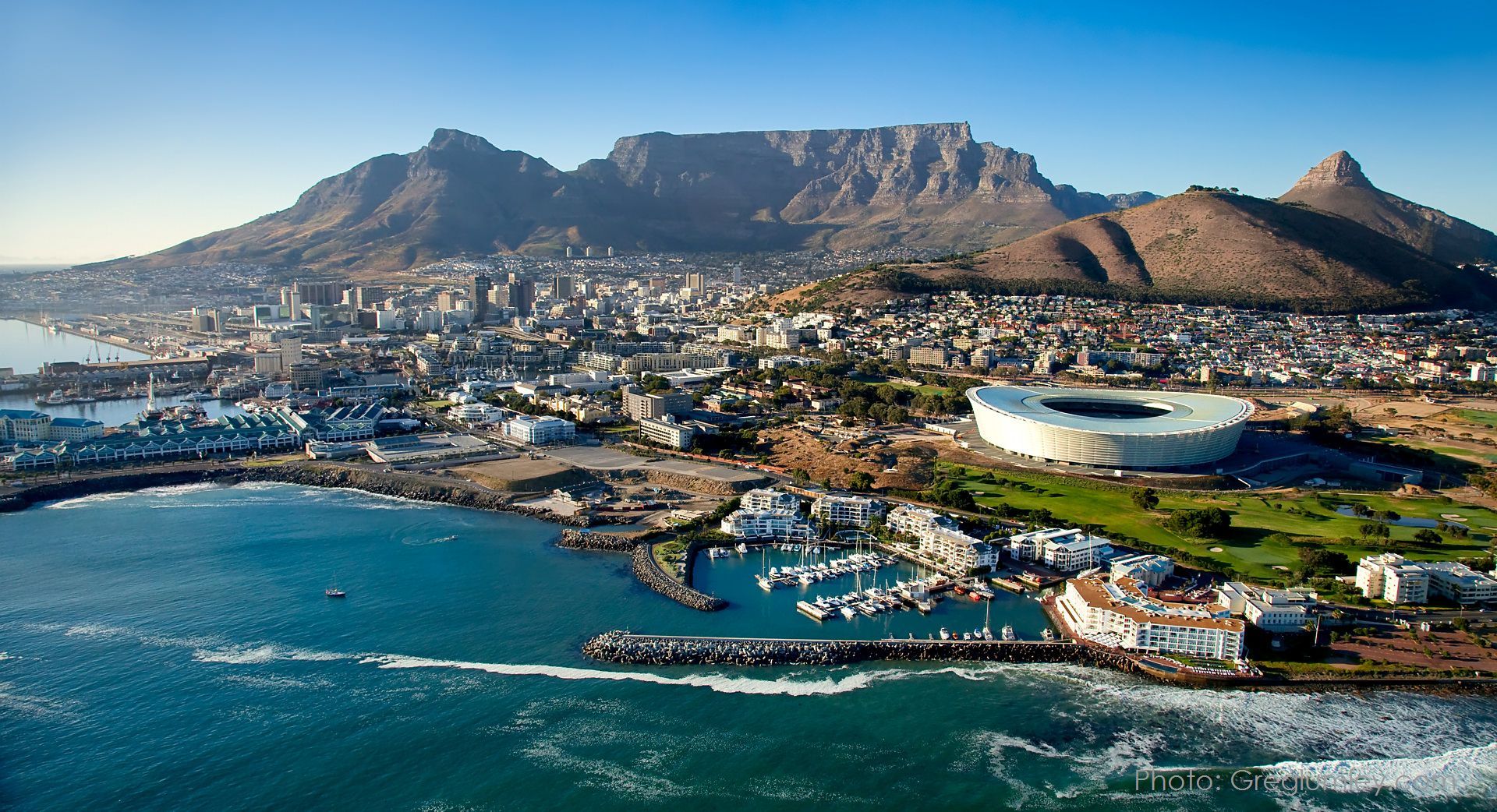 Aerial view of Cape Town, South Africa, with Table Mountain in the background, ocean in the foreground, and stadium.