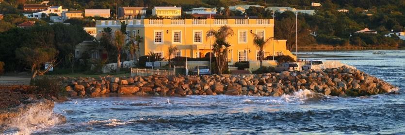 A yellow building sits on a rock breakwater at the edge of the ocean, with a shoreline and buildings in the background.